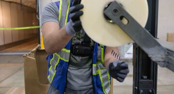 A worker wears a harness while replacing packing tape on a machine.