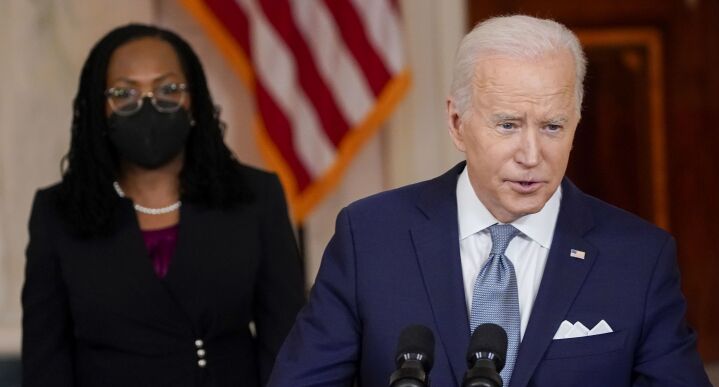 President Joe Biden stands at a lectern as he announces the nomination of Ketanji Brown Jackson to the U.S. Supreme Court. Jackson, who is wearing a mask, stands behind him and looks on.